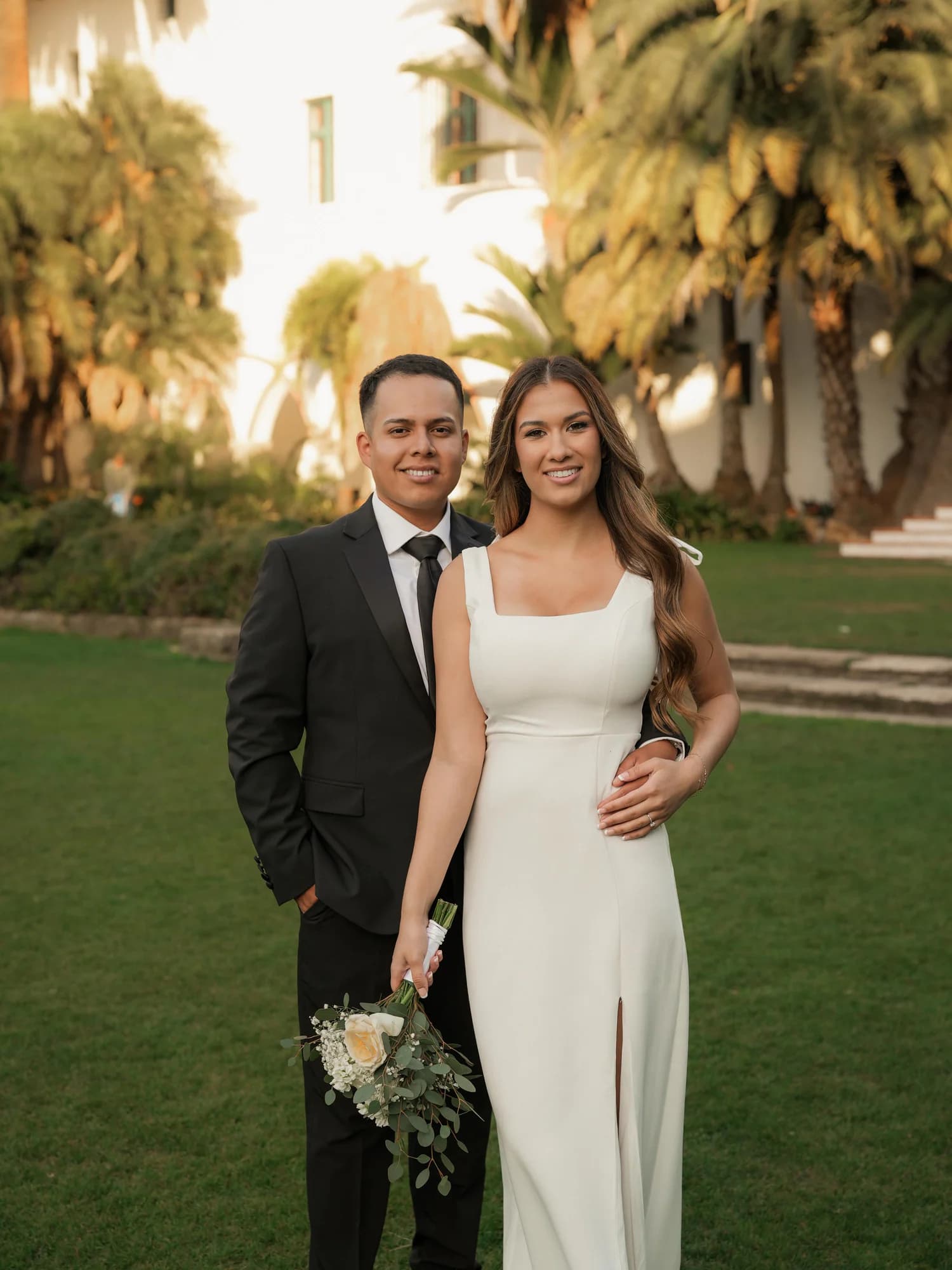 A couple in wedding attire standing together on a palm-lined lawn